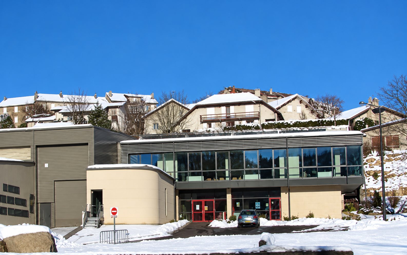 Un bâtiment moderne à grandes baies vitrées et aux murs beige, entouré de neige, avec des maisons de village en hauteur sous un ciel bleu.