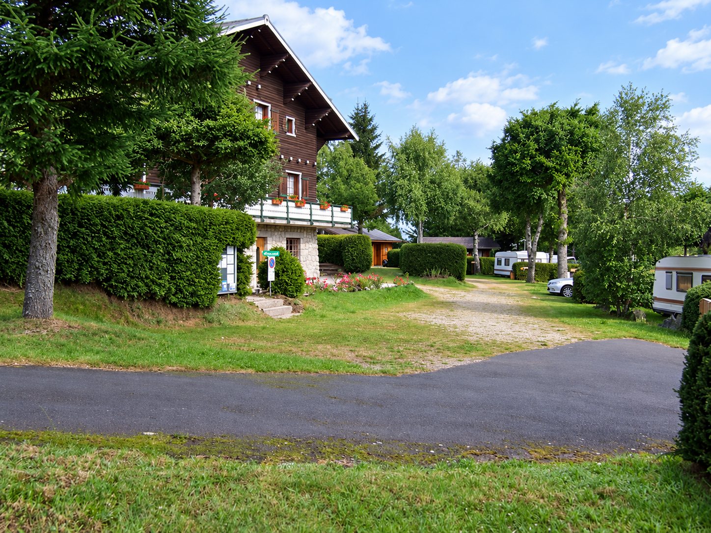 Un chalet en bois avec balcon fleuri domine l'entrée d'un camping bordé de haies et d'arbres, où l'on aperçoit des caravanes.