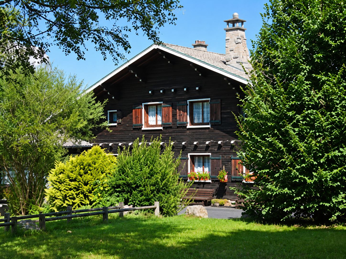 Un grand chalet en bois sombre aux volets clairs se dresse derrière des arbres et des buissons verts, sous un ciel bleu.