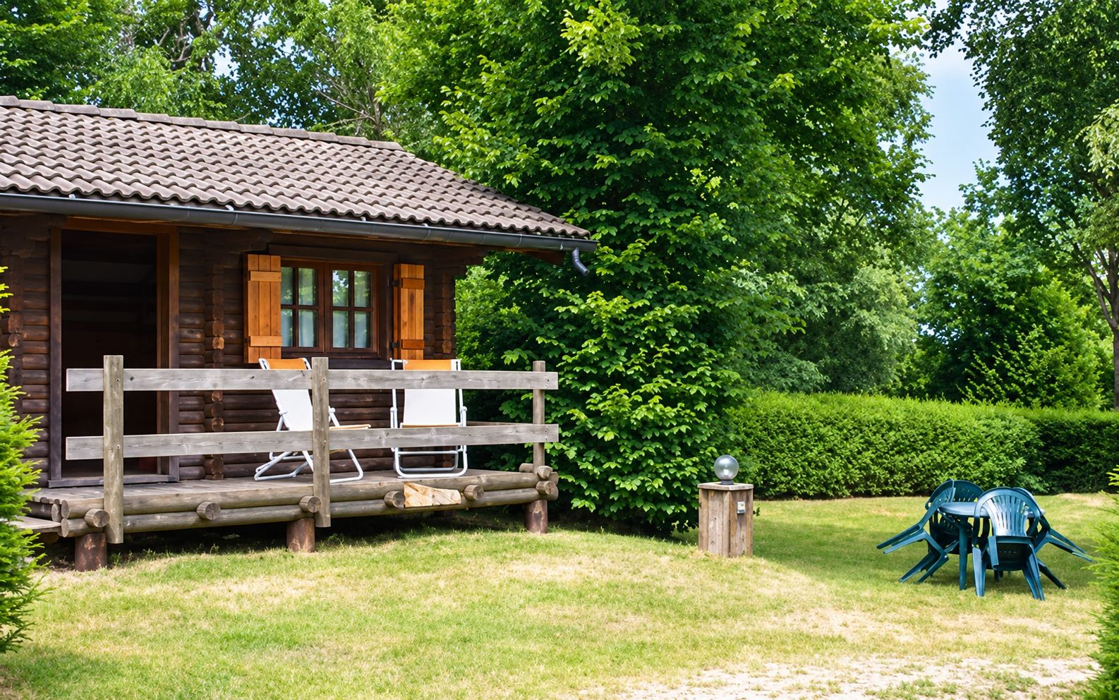 Un petit chalet en bois au toit de tuiles avec une terrasse équipée de chaises longues blanches, entouré d'arbres, d'une haie et d'une pelouse avec un salon de jardin.