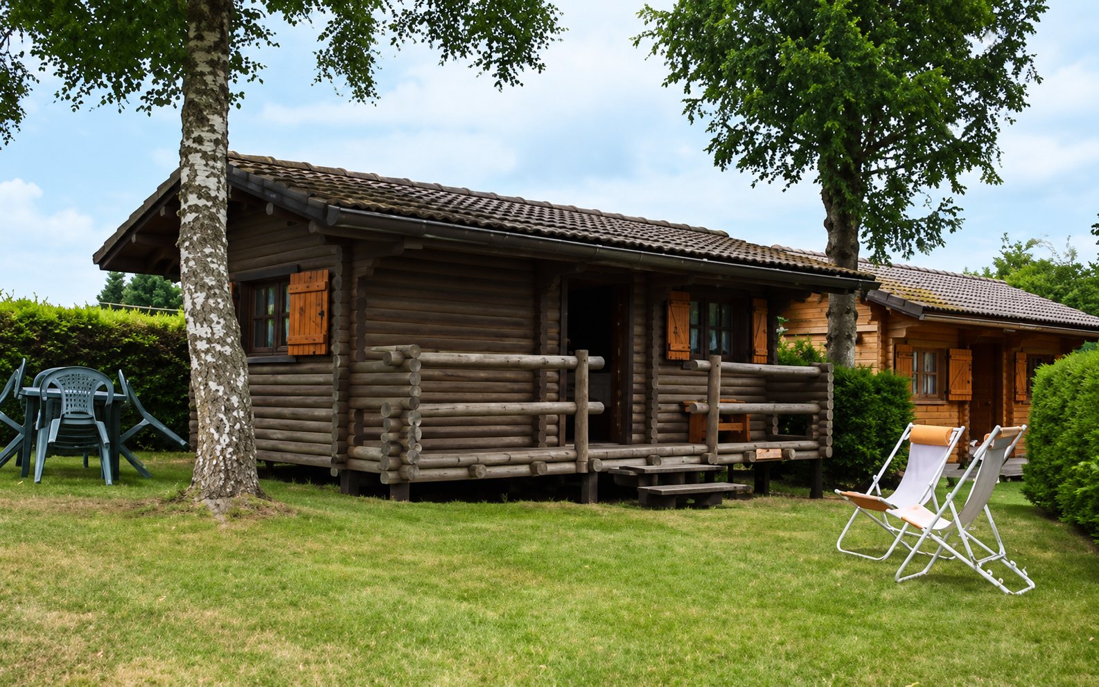 Un chalet en rondins avec volets orange et petite terrasse en bois, entouré d'une pelouse avec une table et des chaises longues sous les arbres.