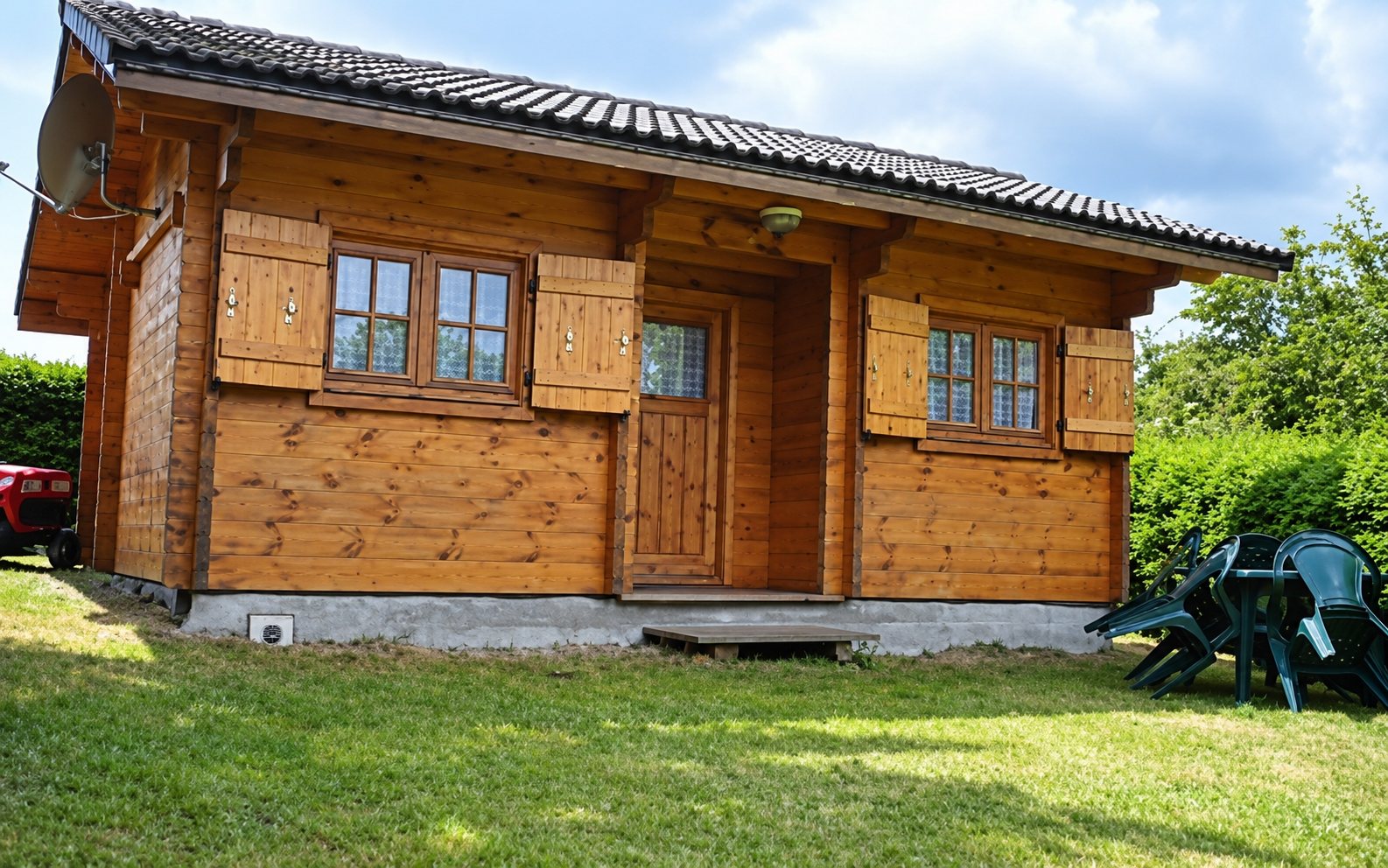 Un petit chalet en bois massif avec des volets en bois et un toit sombre, entouré d'une pelouse et de verdure.