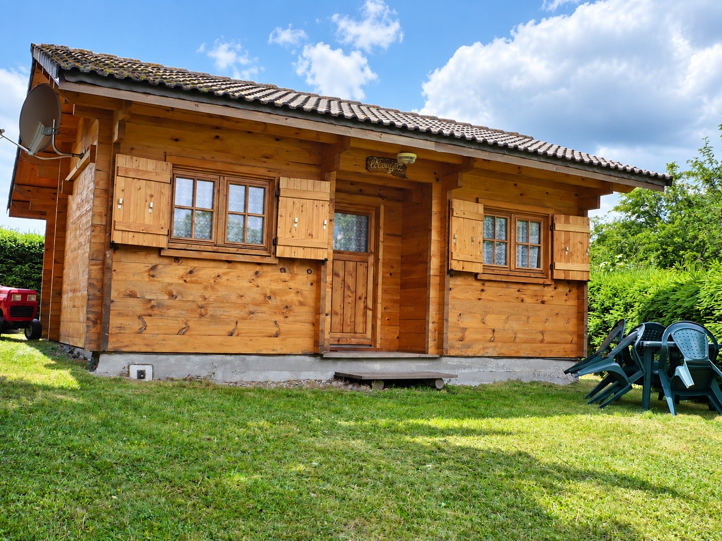 Un petit chalet en bois à toit de tuiles, avec volets ouverts, entouré d'une pelouse et de chaises de jardin vertes empilées à droite.