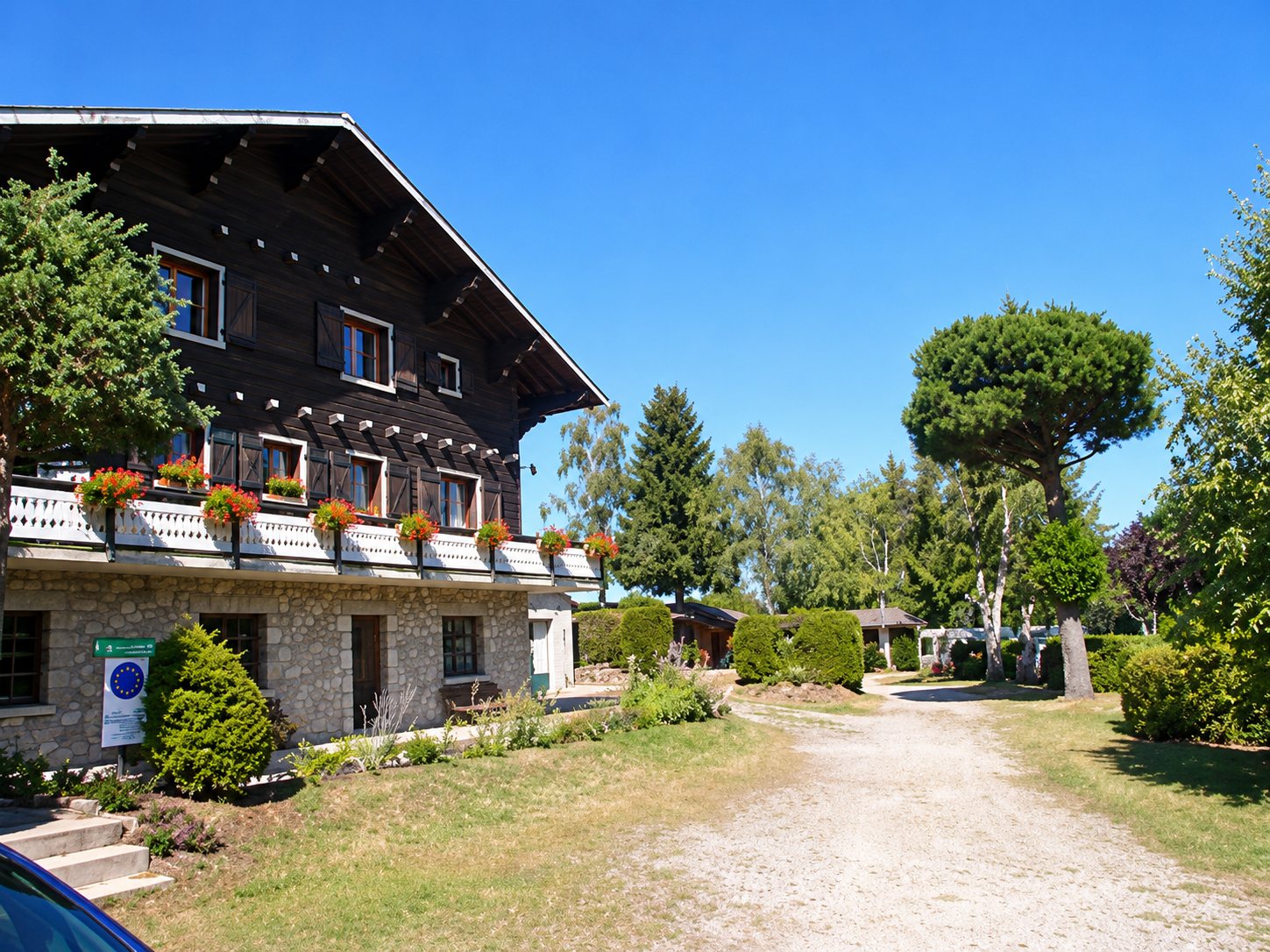 Un grand chalet en bois sombre aux balcons ornés de fleurs borde un chemin de gravier sous un ciel bleu, entouré d'arbres et de pelouse.