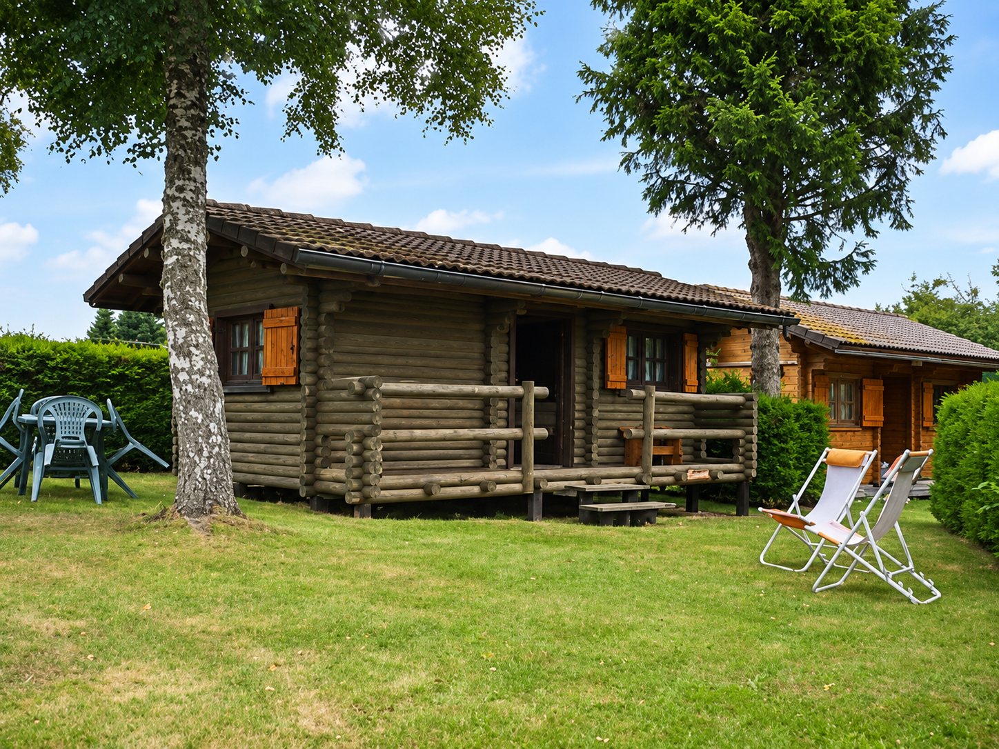Un chalet en rondins de bois aux volets orange, avec une terrasse, entouré d'une pelouse où se trouvent des chaises longues et une table de jardin.