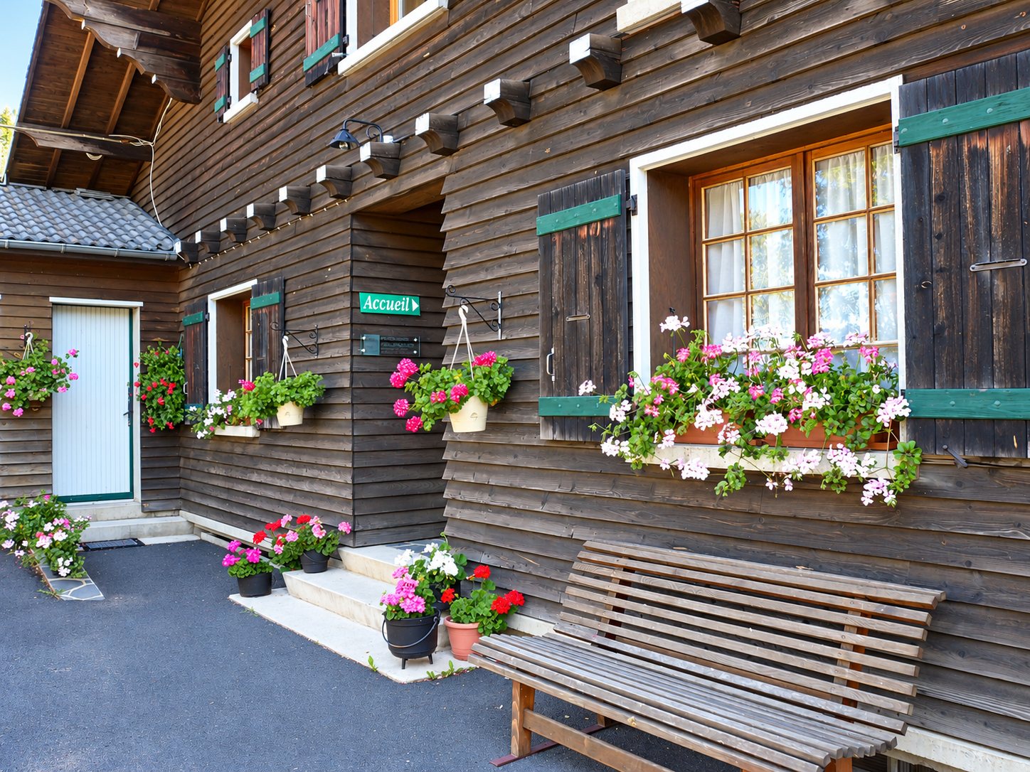 Façade d'un chalet en bois sombre aux volets verts, ornée de géraniums en pots et en jardinières, avec un banc en bois et un panneau vert indiquant l'accueil.