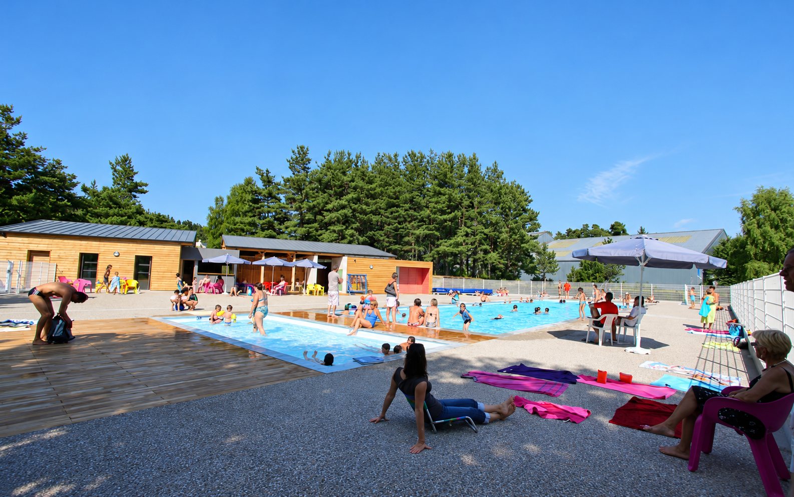 Des familles se baignent et se détendent autour d'une piscine extérieure par une journée ensoleillée, avec des bâtiments en bois et des pins en arrière-plan.