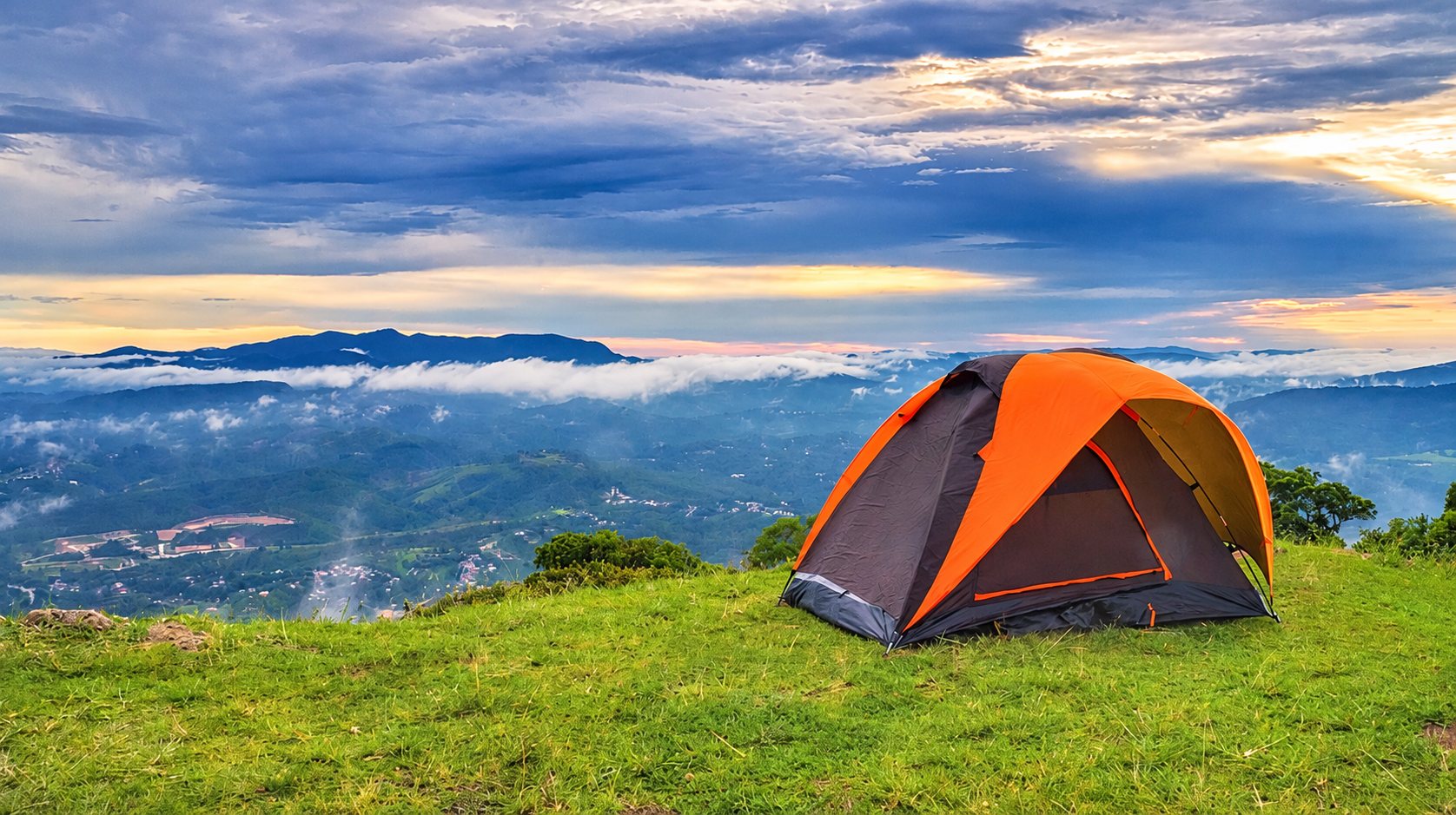 Une tente de camping orange et grise est installée sur une colline verdoyante qui domine une vallée recouverte de brume sous un ciel nuageux.